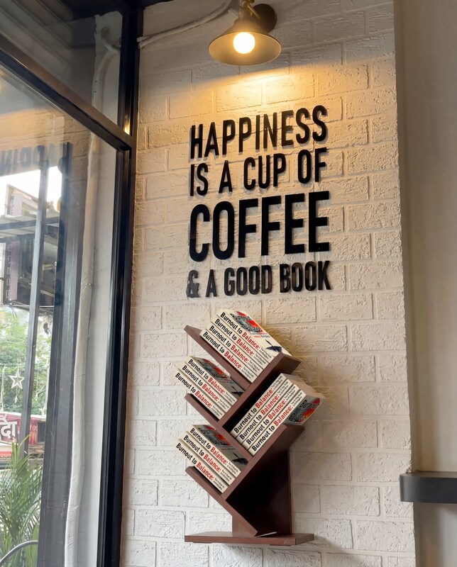 Wall art in a coffee shop reading Happiness is a Cup of Coffee and a Good Book, with a display of Burnout to Balance books by Andrea Gomez